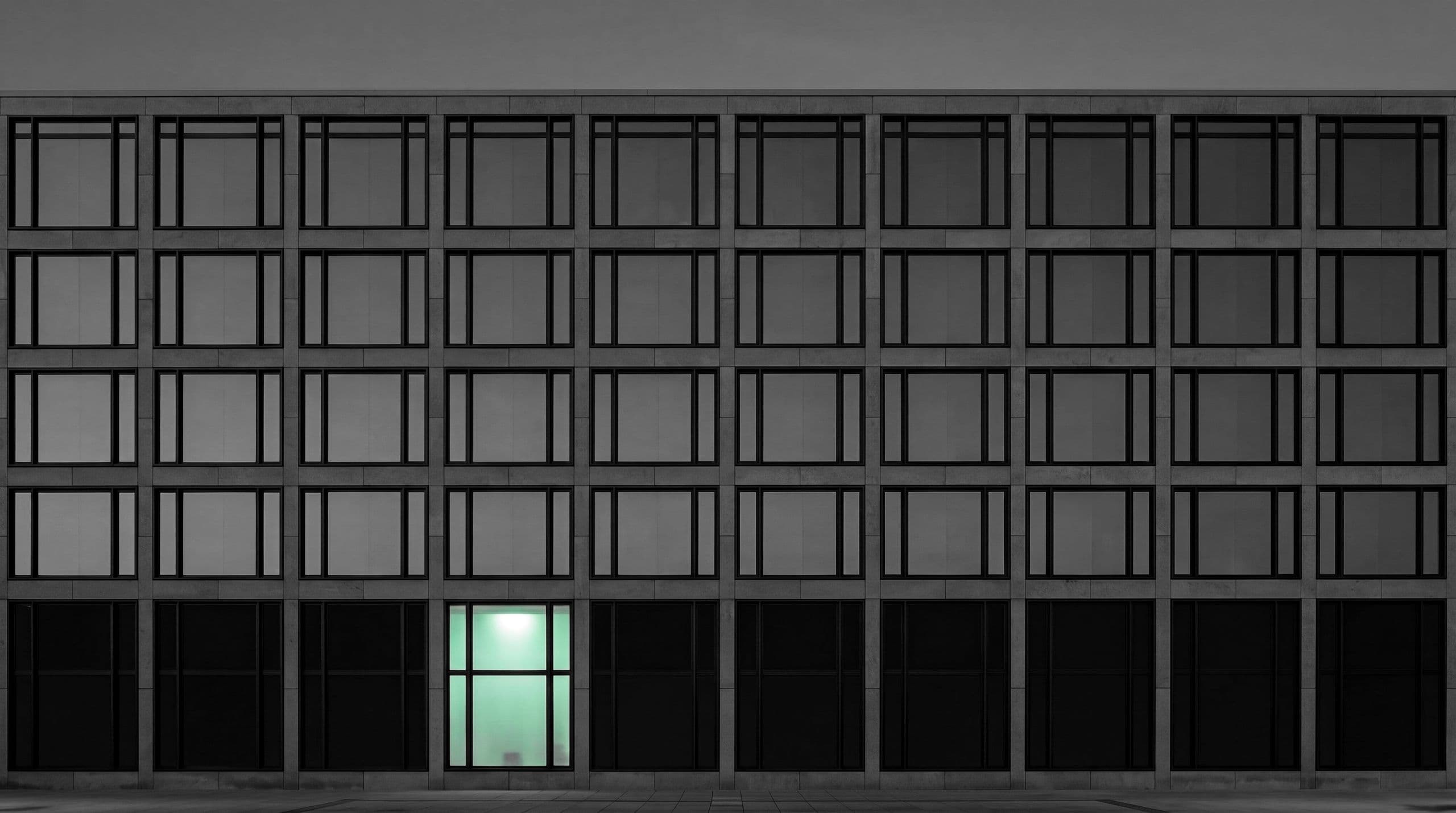 Una fachada de oficinas en blanco y negro al atardecer con una sola ventana encendida en verde menta